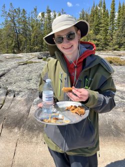 Person outdoors holding a tray with fried food and a water bottle, wearing sunglasses, a hat, and a green jacket.
