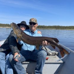 Two people sitting in a boat on a lake, holding a large fish together and smiling at the camera.