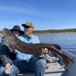 Two people in a boat hold a large fish over their laps, with a lake and treeline visible in the background.