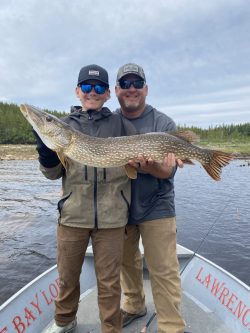 Two people on a boat hold a large northern pike fish in front of a river and forested shoreline.