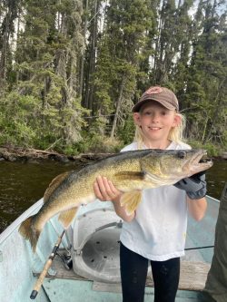 Young person on a boat holding a large fish, with trees and water visible in the background.