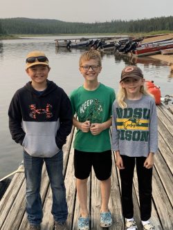 Three kids stand on a wooden dock by a lake with boats and forest in the background, smiling at the camera.