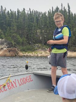 A boy in a life jacket stands on a boat holding a fishing rod with a fish on the line near a forested shoreline.