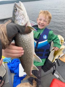 A smiling child in a life jacket is on a boat as an adult holds up a freshly caught fish in the foreground.