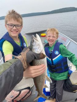 Two children in life jackets smile on a boat as an adult holds up a freshly caught fish in the foreground.