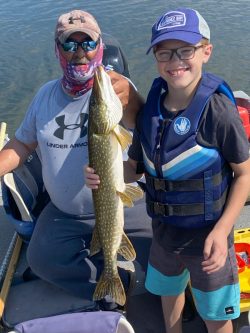 An adult and a child on a boat hold a large fish, both smiling; the child wears a life jacket and cap.