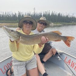 Two people in a boat display a large fish they caught, with a forested shoreline in the background.