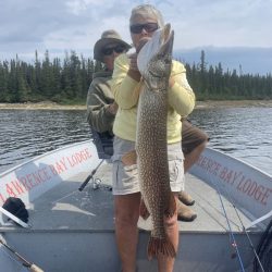 Two people in a boat on a lake, with one person holding a large fish vertically in front of them.