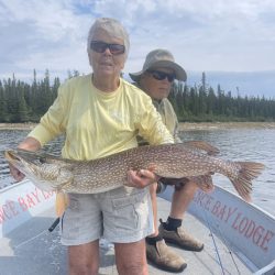 Two people on a boat, one holding a large fish with "Lodge" visible on the side and trees in the background.