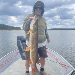 Person in a hoodie and sunglasses holding a large fish on a boat at Lawrence Bay Lodge, with water and trees in the background.