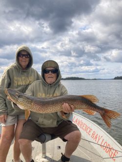 Two people in hoodies on a boat hold a large fish, with a lake and cloudy sky in the background.