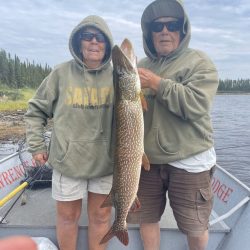 Two people in hoodies hold a large fish on a boat by a lake, with trees and cloudy sky in the background.