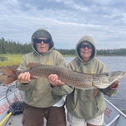 Two people in hoodies and sunglasses hold a large northern pike fish on a boat by a lake with trees in the background.