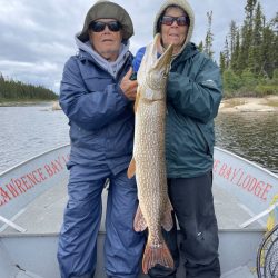 Two people in outdoor gear hold a large fish on a boat, with trees and water in the background.
