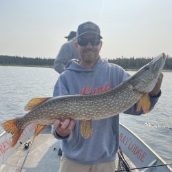 Man standing on a fishing boat holding a large northern pike with water and trees in the background.