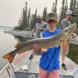 A person on a boat holds a large fish with two others fishing in the background by a forested shoreline.