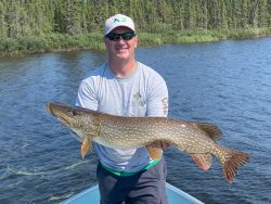 Man standing on a boat holds a large northern pike over a lake with trees in the background.