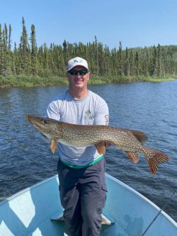 Man standing on a boat holding a large fish with a forest and lake in the background.