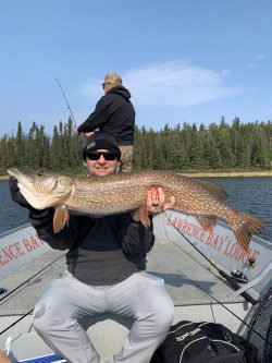A man sitting on a boat holds a large fish, while another person stands fishing in the background on a lake.