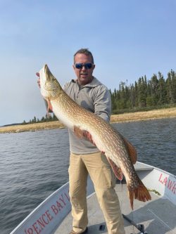 Man standing on a boat holding a large fish with water and trees visible in the background.
