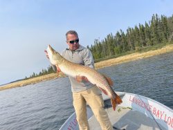 A man stands on a boat holding a large fish with a forested shoreline in the background.