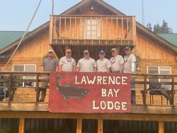 Six men stand on the deck of a wooden lodge with a large sign reading "Lawrence Bay Lodge" and a fish illustration.