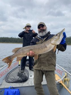 Man holding a large fish on a boat, while another person takes a photo; lake and forest in the background.