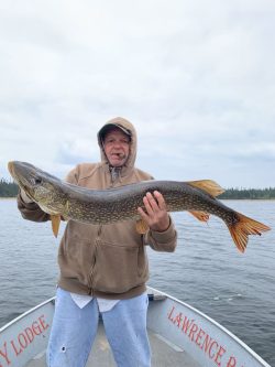 Person in a brown hoodie holding a large fish on a boat, with water and trees visible in the background.