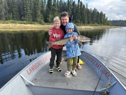 An adult and two children stand on a boat holding a large fish, with trees and water in the background.