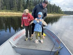 Two children and an adult stand on a boat; the child in front holds a large fish. The boat reads "Lawrence Bay Lodge.