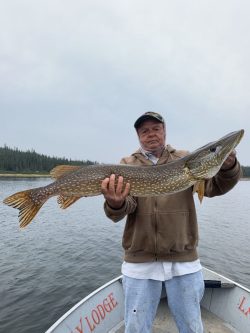 A person in a brown jacket stands on a boat holding a large fish over a lake with trees in the background.