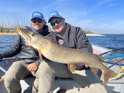 Two people sitting in a boat hold a large northern pike fish; water and shoreline appear in the background.