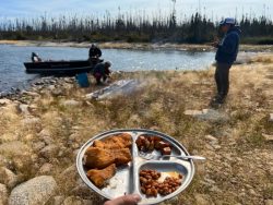 A metal plate with fried fish, beans, and potatoes is held by a lake, with people and a boat in the background.