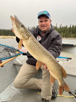 A man on a boat holds a large northern pike fish with a forested shoreline in the background.