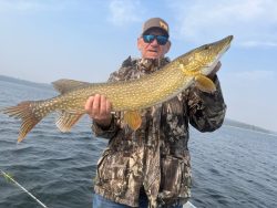 Person wearing camouflage jacket and sunglasses holds a large fish on a boat over a lake under a cloudy sky.