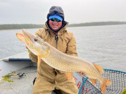 Person in waterproof clothing holds a large fish on a boat in a lake on a cloudy day, with a forest in the background.