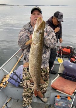 Man on a boat holding a large fish, with another person in the background preparing fishing gear.