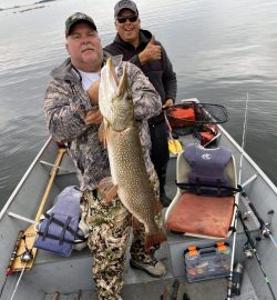 Two men in a boat, one holding a large fish, the other giving a thumbs up, fishing gear visible around them.