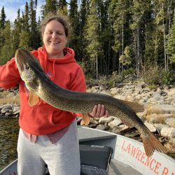 Person in a red hoodie standing in a boat holding a large fish with trees and rocks in the background.