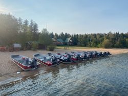 A row of small motorboats is lined up on a sandy shore with cabins and trees in the background.
