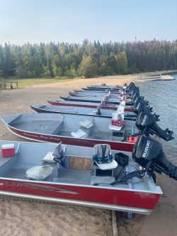 A row of red motorboats with life jackets lined up on a sandy shore next to a lake and forested background.