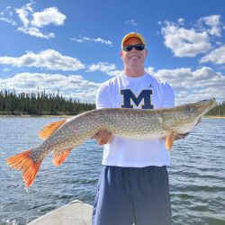 Man wearing a Michigan shirt holding a large fish on a boat, with water and trees in the background under a blue sky.