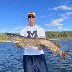 Man in Michigan shirt and cap holding a large fish on a boat, with lake and forest in the background.