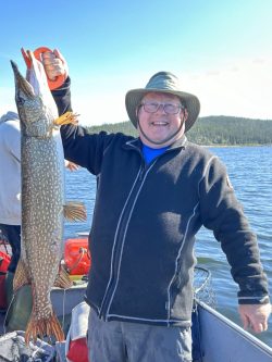 Smiling person in a hat holds up a large fish on a boat, with a lake and forest in the background.