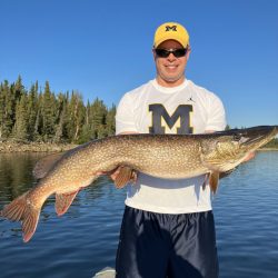 Man wearing a yellow cap holds a large fish on a lake, with trees and blue sky in the background.