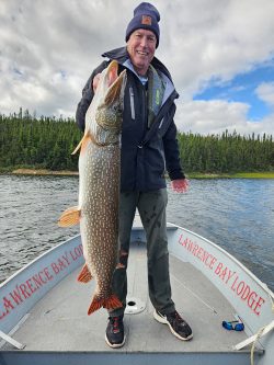 A person standing on a boat holds a large fish with a forest and cloudy sky in the background.
