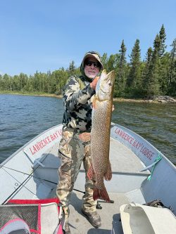 Person in camouflage clothing holds a large fish while standing on a boat in a lake with trees in the background.