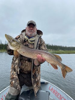 A man in camouflage gear stands on a boat holding a large fish over the water on a cloudy day.