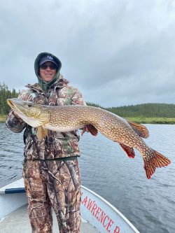 Person in camouflage clothing holding a large fish on a boat, with water and forested shoreline in the background.