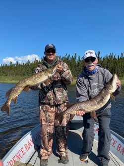 Two men on a boat holding large fish they caught, with trees and blue sky in the background.